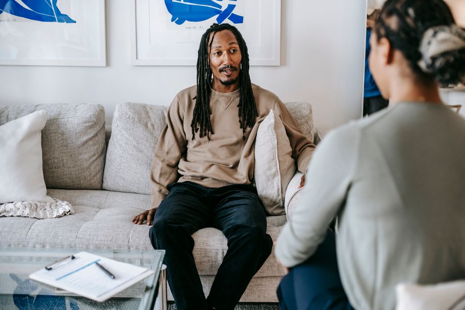 African American male client with dreadlocks sitting on sofa and speaking with unrecognizable female therapist during psychotherapy session in cozy room