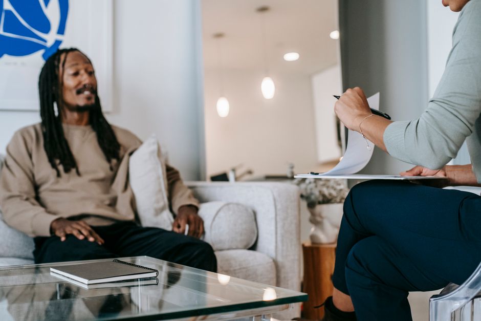Thérapie avec partenaire de substitution : ce que le cadre change et ce qu’il ne prouve pas African American male with dreadlocks sitting on sofa in front of unrecognizable female therapist with clipboard during psychotherapy appointment in office
