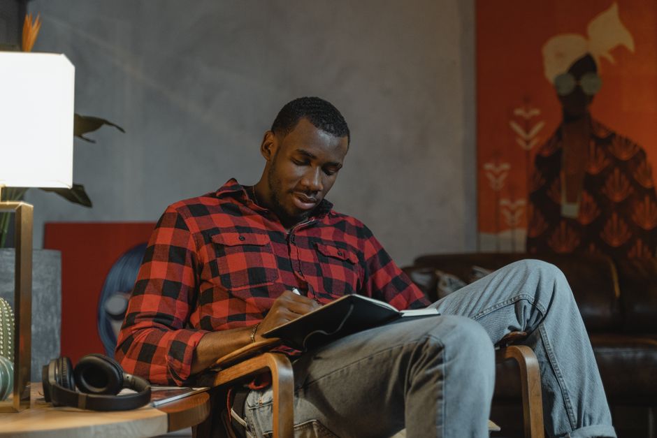 Sublimation en psychologie : transformer une impulsion sans se mentir African American man in a checkered shirt writes in a notebook while sitting indoors.