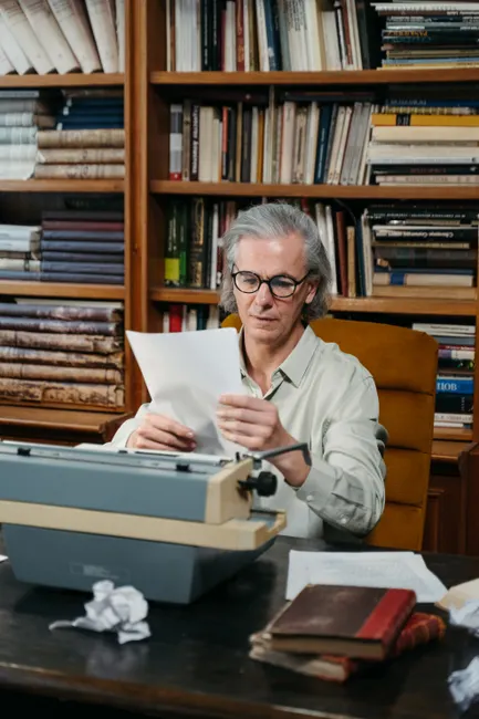 An elderly man reading papers inside a library, sitting with a typewriter.