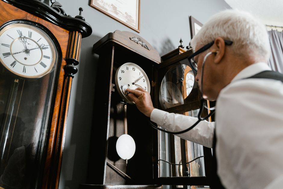 An elderly man with white hair uses a stethoscope to check and adjust a vintage wall clock indoors.