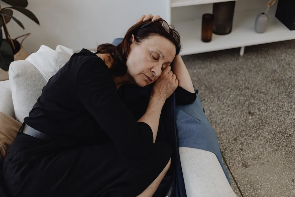 An elderly woman rests peacefully on a sofa in a cozy domestic setting, wearing eyeglasses.