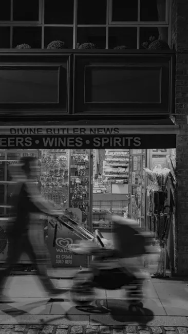 Black and white image capturing a person with stroller walking past a store at night.