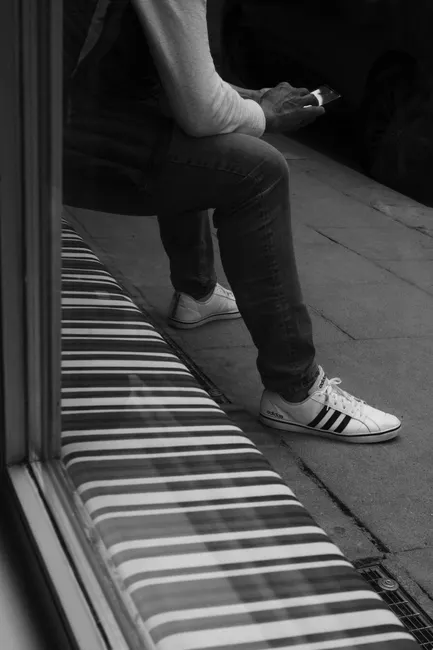 Black and white photo of a person sitting on a bench using a smartphone outdoors.
