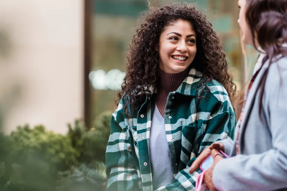 Cheerful woman with long curly hair in casual clothes standing and communicating with friend on street in daylight