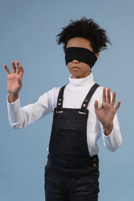 Child with afro hairstyle in denim overalls blindfolded, exploring with hands.