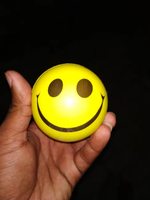 Close-up of a hand holding a yellow smiley face stress ball against a dark background.