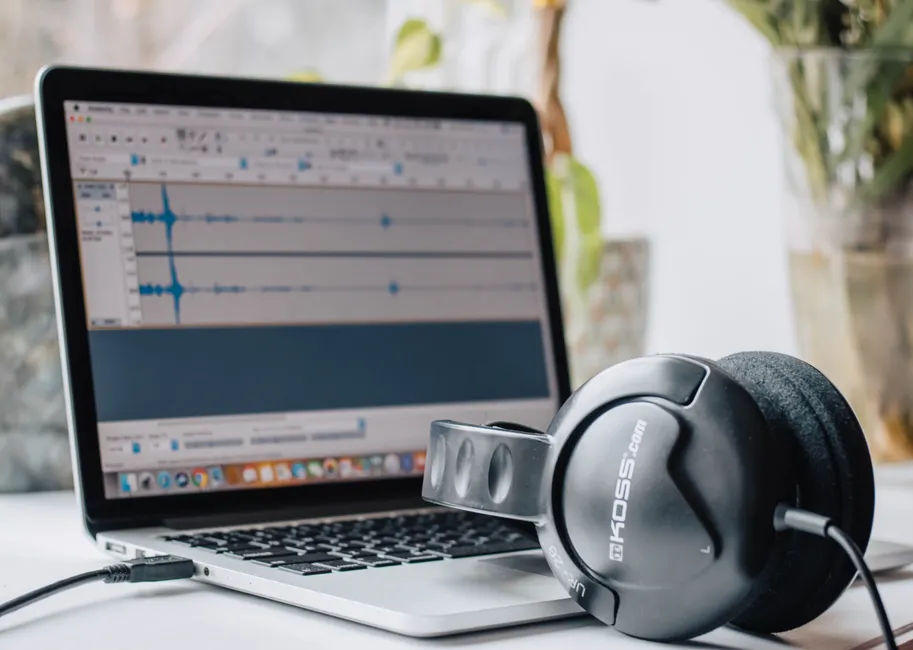 Close-up of a laptop with audio editing software and headphones in a home office setting.