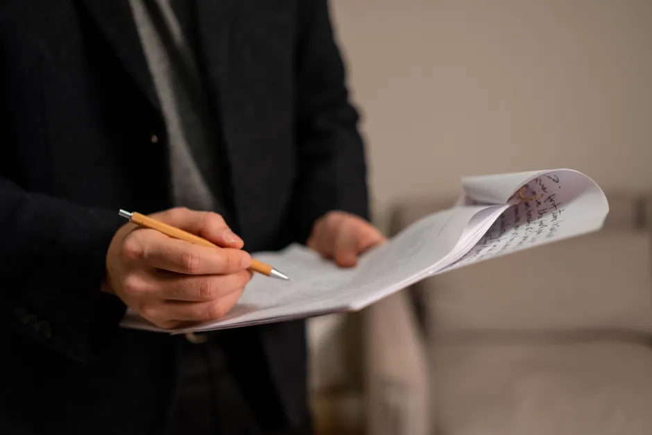 Close-up of a man holding and reviewing handwritten papers with a pen indoors.