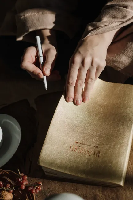 Close-up of hands writing in a gold-covered notebook with a pen on a wooden table.