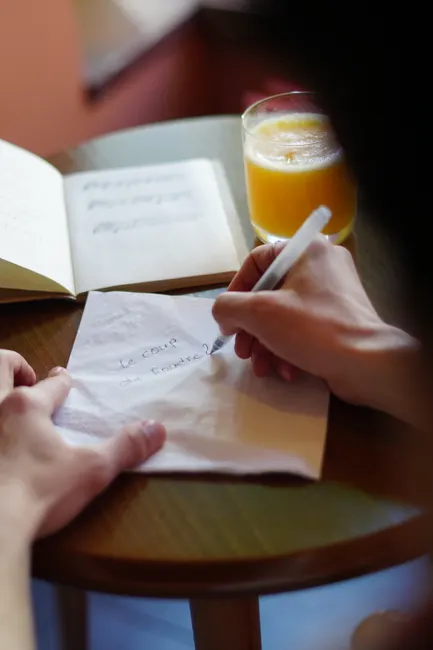 Close-up of hands writing on a napkin with a pen, alongside a notebook and juice glass.