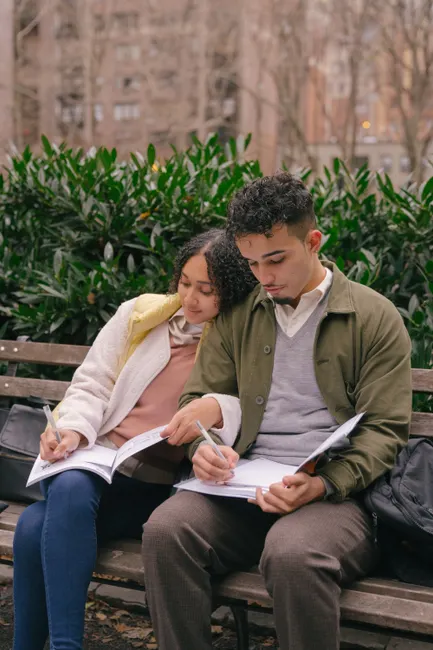 Concentrated ethnic curly couple in casual outfit sitting on bench together and writing in workbooks while preparing homework in city park