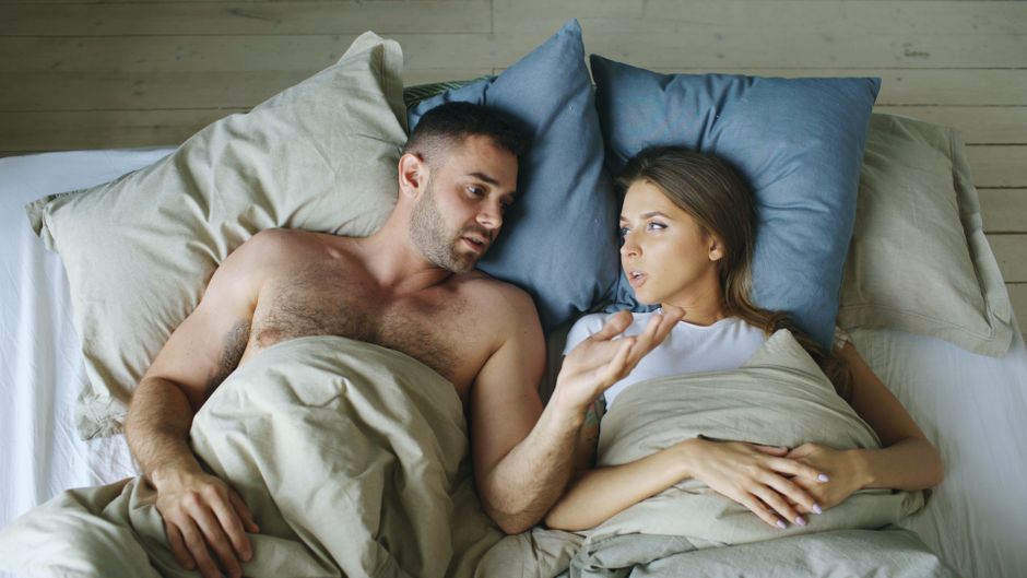 Éjaculation retardée : comprendre les causes et sortir de la pression Couple lying in bed under blankets, engaged in conversation.