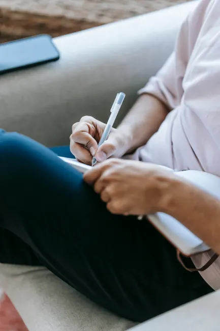 Crop anonymous person in casual clothes taking notes in copybook while sitting in comfy armchair