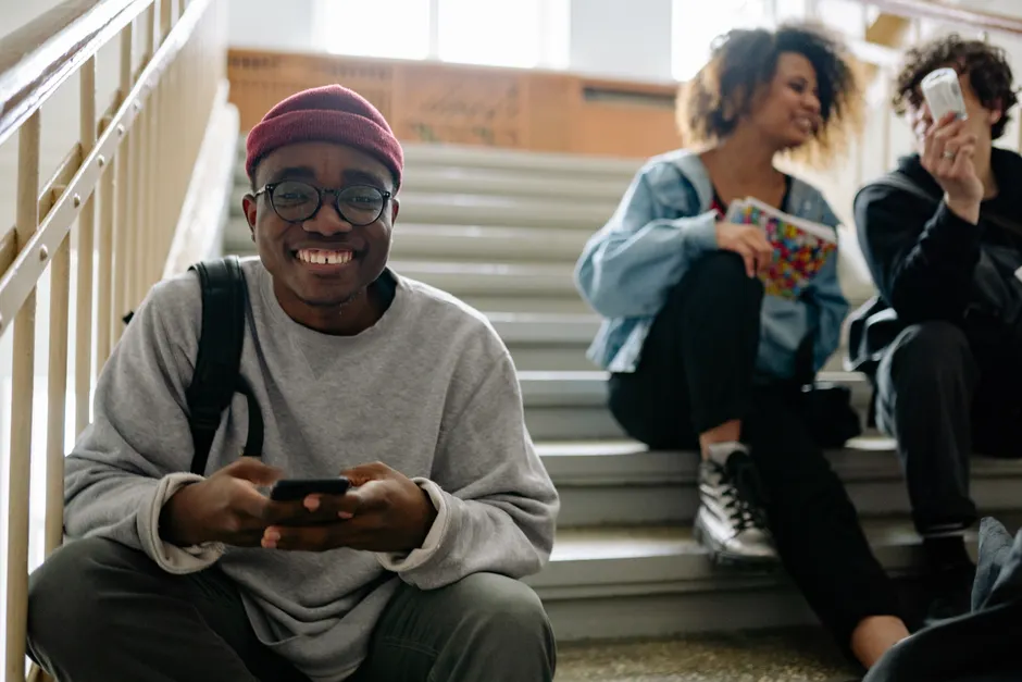 Diverse group of happy students sitting on school staircase, smiling and using mobile phones.