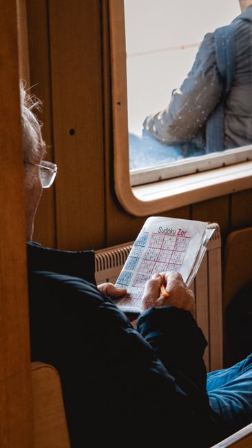 Elderly man enjoys a relaxing moment solving Sudoku puzzle while sitting by a window indoors.