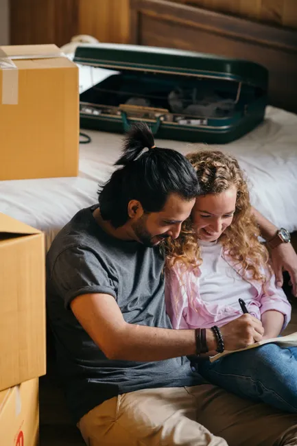 Ethnic man and smiling curly woman hugging while sitting on floor in bedroom and planning future with notebook and pen in new apartment among carton boxes and cases