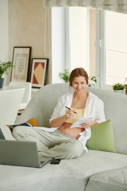 Expecting mother seated on sofa, writing in journal with a happy expression. Indoor setting.