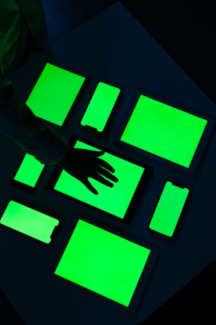 Hand reaching over various electronic devices with glowing green screens, emphasizing digital connectivity.