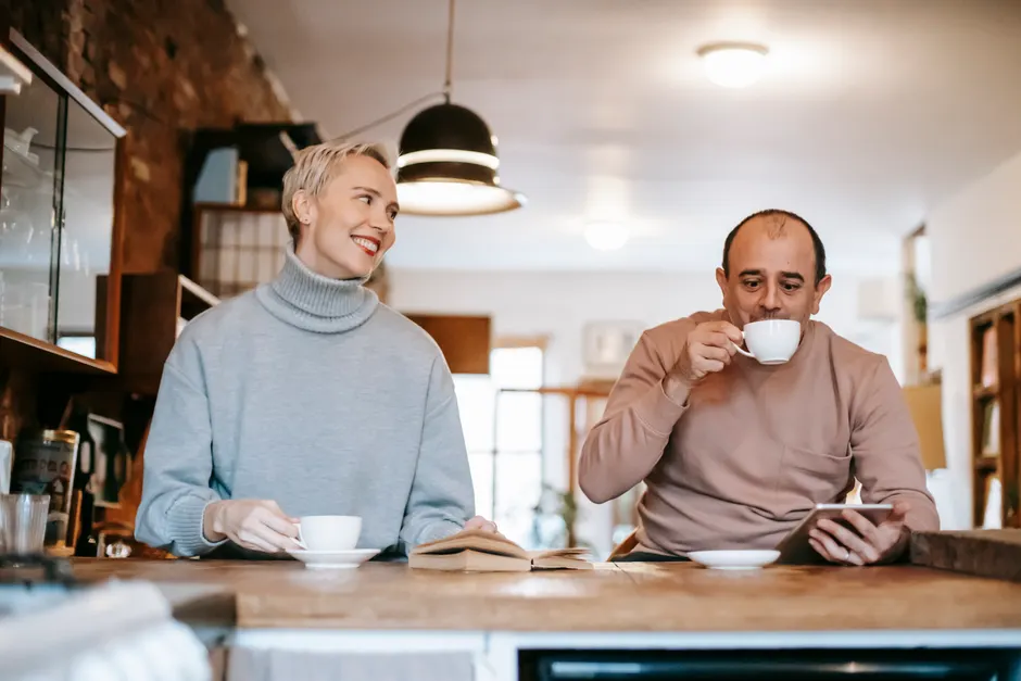 Happy adult couple at counter drinking tea from cups with saucer near opened book and while using tablet in light room
