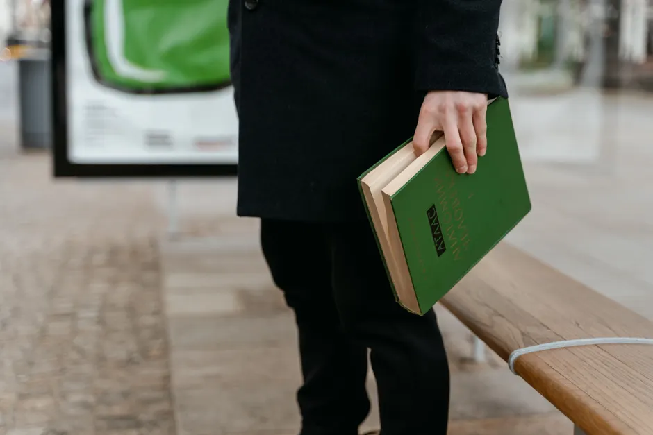 Partial view of person holding green book while standing near a wooden bench outdoors.