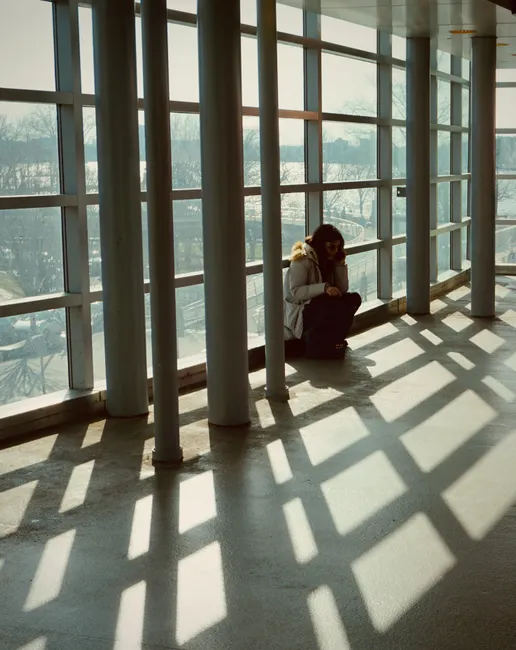 Person sitting alone by large windows casting shadows indoors.
