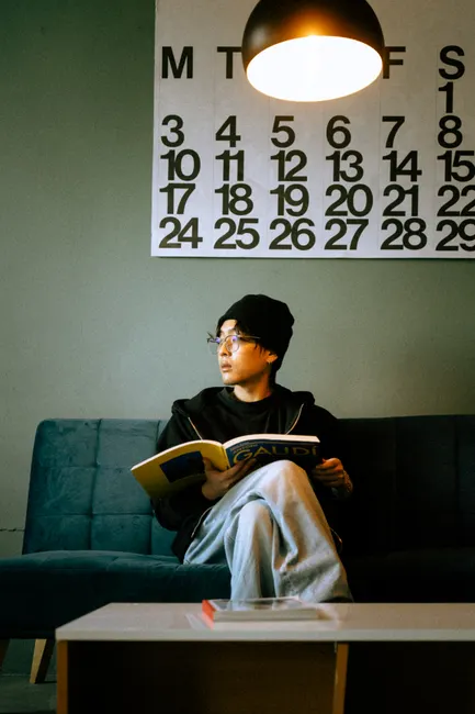 Person wearing glasses reads a book on a sofa with large calendar art on wall.