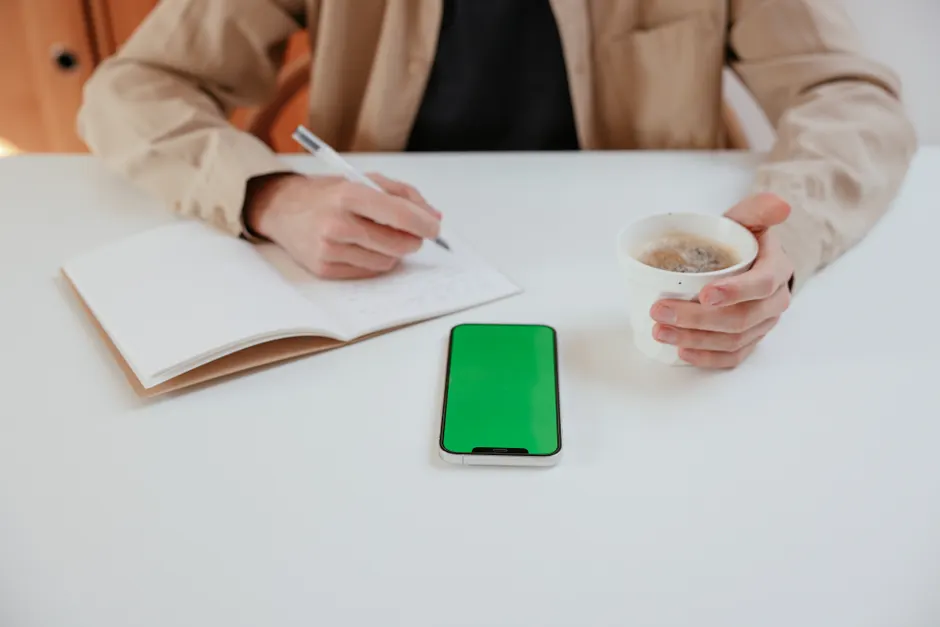 Person writing in notebook while holding coffee and smartphone with green screen on table.