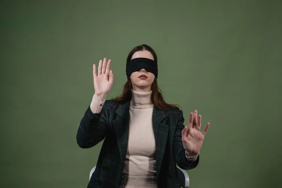 Portrait of a woman blindfolded with raised hands against a green background suggesting sensory exploration.