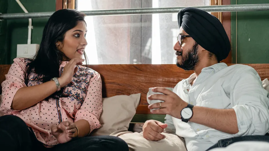 Positive happy Sikh couple wearing casual clothes and turban having lively conversation while resting on comfortable bed with cup of hot drink