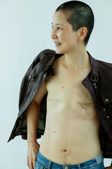 Positive mature ethnic female in jacket and jeans standing with scars on chest after breast removal surgery from cancer in light studio on white background