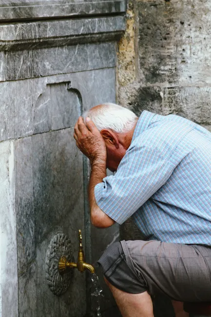 Senior man in casual attire washes face at an outdoor tap, emphasizing refreshment and daily life.