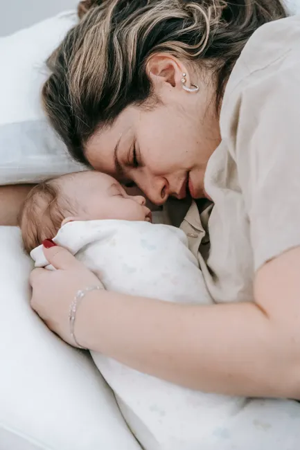 Side view of caring mother with closed eyes embracing cute sleeping infant baby while lying on comfortable bed in bedroom