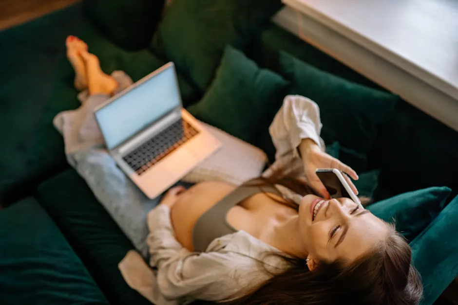 Smiling pregnant woman lounging on sofa, talking on a smartphone with a laptop nearby, enjoying a cozy indoor moment.