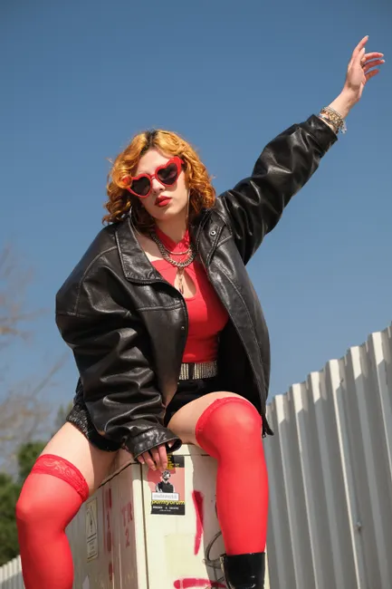 Stylish woman wearing a leather jacket, red accents, and heart-shaped sunglasses posing outdoors.