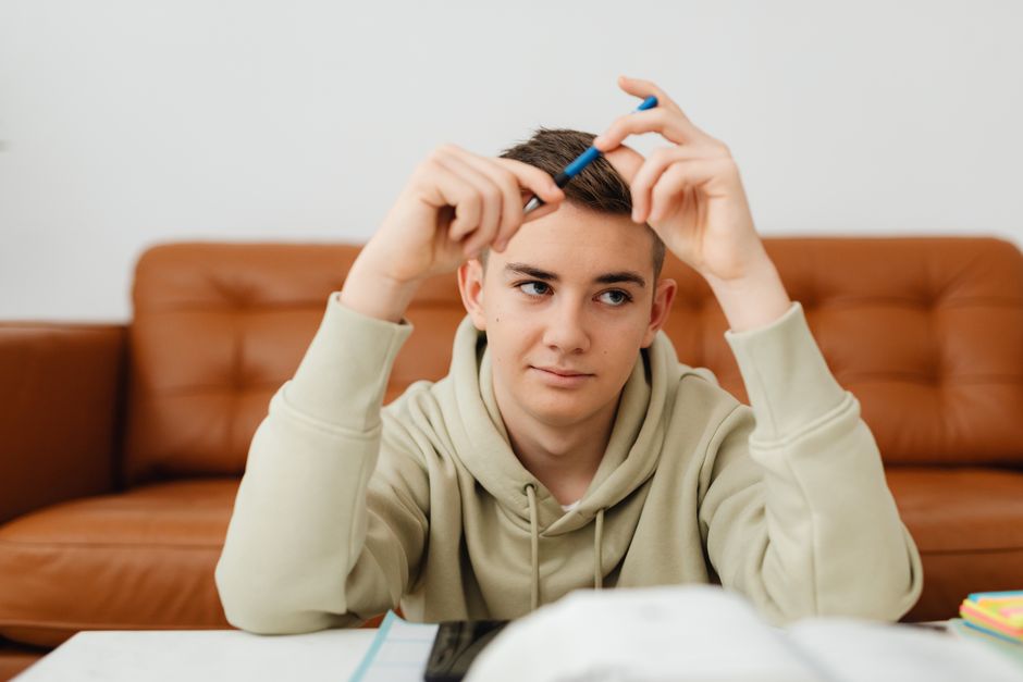 Teenager in hoodie contemplating with pen while studying on a brown sofa indoors.