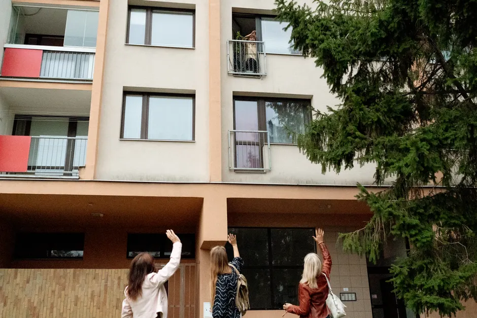 Three women waving to an elderly woman on an apartment balcony in Prague.