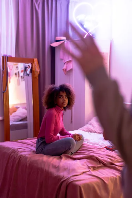 Trendy teenager sitting on bed in fashionable bedroom with neon light ambiance.
