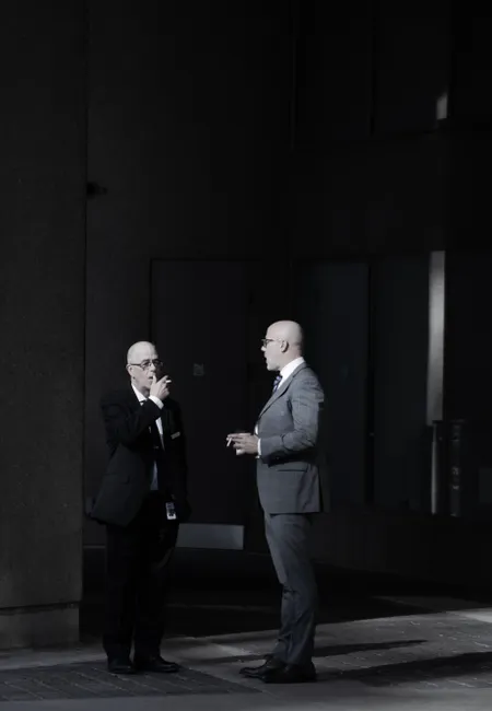 Two business professionals in suits having a conversation and smoking outside an urban building.