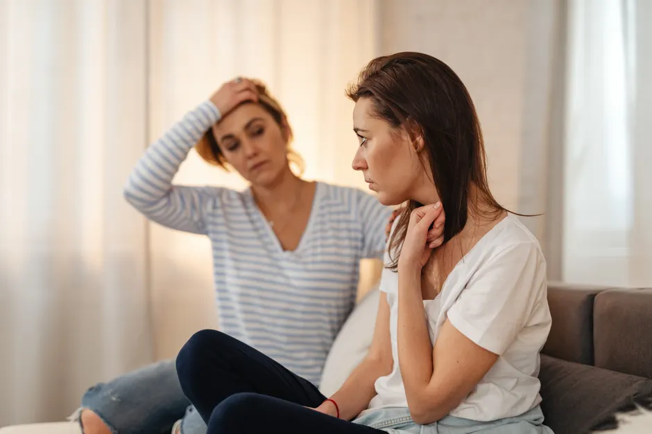 Two women sitting on a sofa showing concern and support for each other indoors.