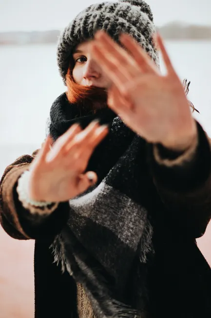Young woman outdoors in winter wearing a knitted cap and scarf, playfully covering her face with hands.