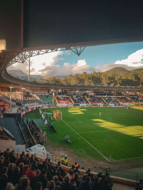 A bustling soccer stadium at sunset with spectators enjoying the game amidst scenic mountains.