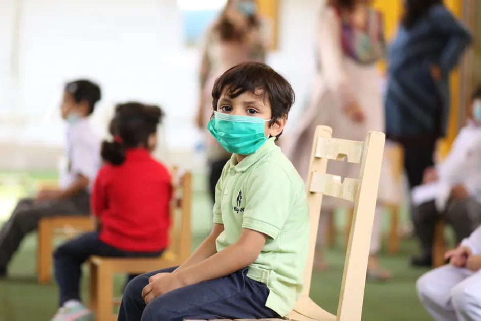 A child wearing a face mask sits on a wooden chair in a socially distanced classroom setting.