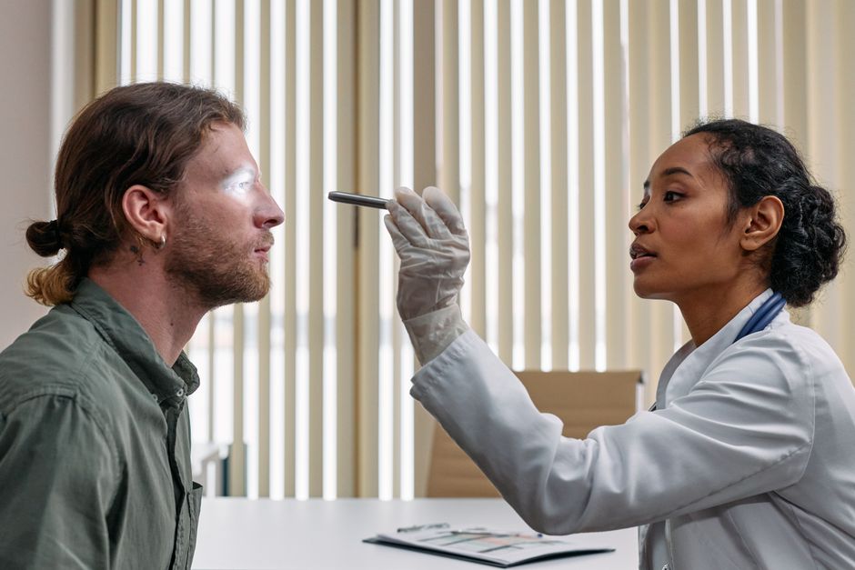 A doctor performs an eye examination on a patient using a flashlight in a clinical setting.