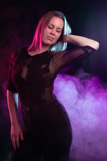 A female model posing confidently in a studio with vibrant neon lights and smoke.
