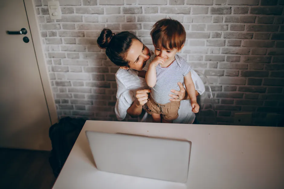 A mother lovingly interacts with her toddler while working on a laptop in a cozy home setting.