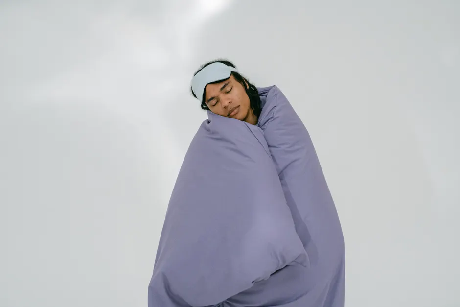 Insomnie : ce qu’un questionnaire repère et ce qu’il ne diagnostique pas A person peacefully sleeping with an eye mask, wrapped in a purple blanket on a white background.