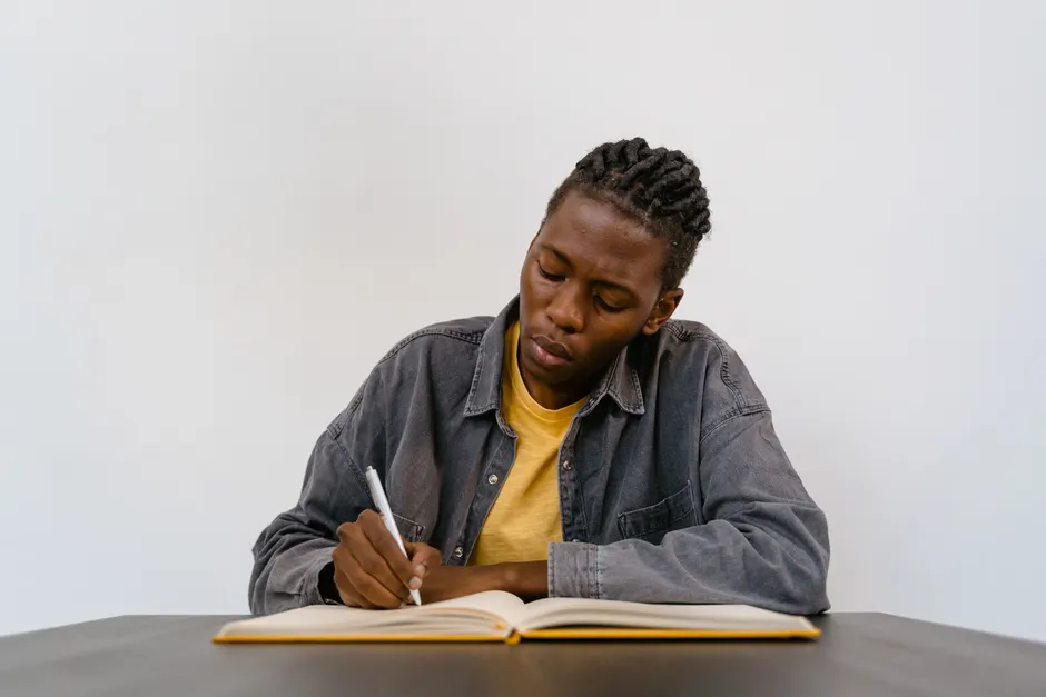 A person with dreadlocks intensely writes in a notebook at a table indoors.
