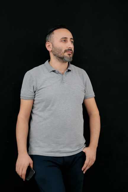 A relaxed man in a grey polo shirt stands against a black background, holding a smartphone.