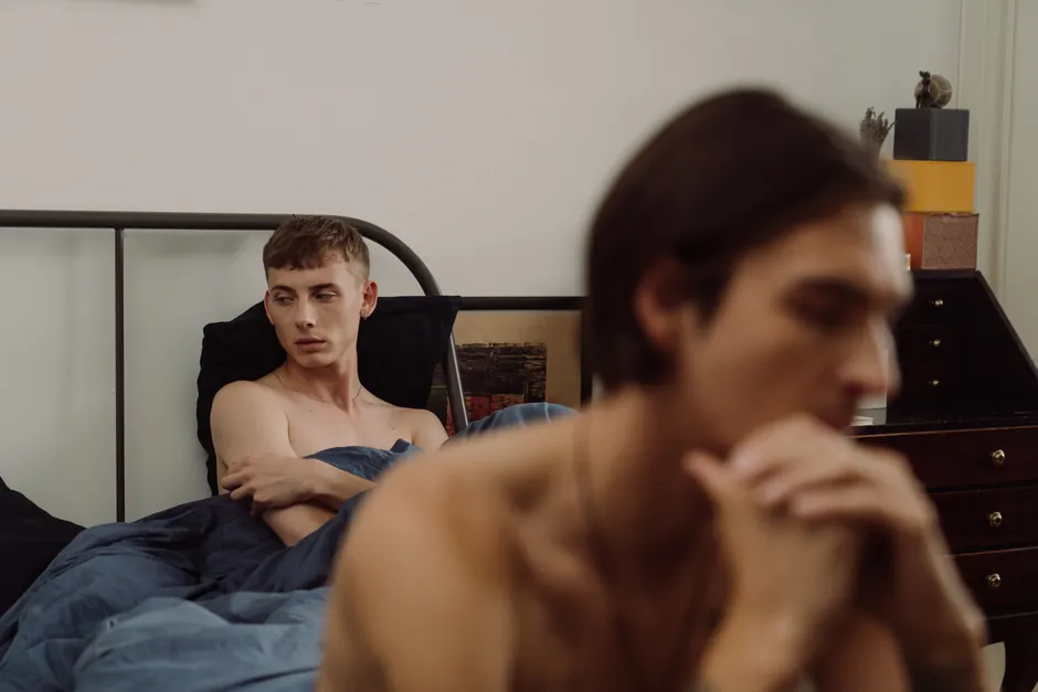 A shirtless couple appears upset in a bedroom, conveying relationship conflict.